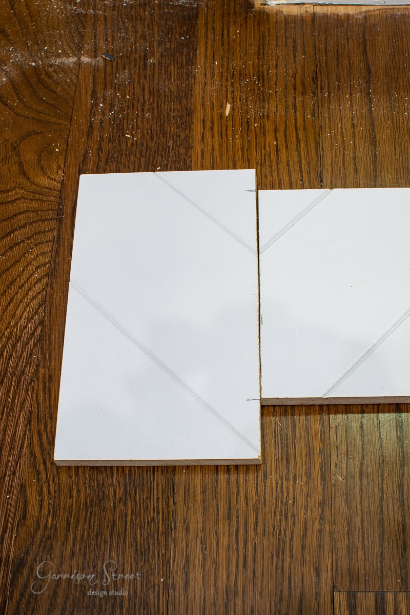 Two white rectangular tiles laid on a wooden floor. The left tile overlaps the right slightly, with visible pencil markings indicating cut lines, resembling stair treads. The wood beneath has a rich brown grain pattern.