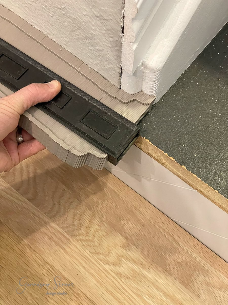 A person uses a contour gauge to measure the edges and contours of stair treads and the white wall-to-dark floor transition. The gauge is pressed against the corner to accurately capture the shape, ensuring each riser aligns seamlessly with its adjacent surface.