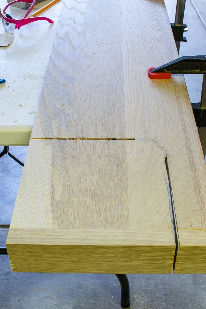 Close-up of a wooden board with two cuts and a marked curved line, held in place by a clamp on a table. Safety goggles and tools are visible nearby, suggesting a woodworking project involving stair treads or risers.