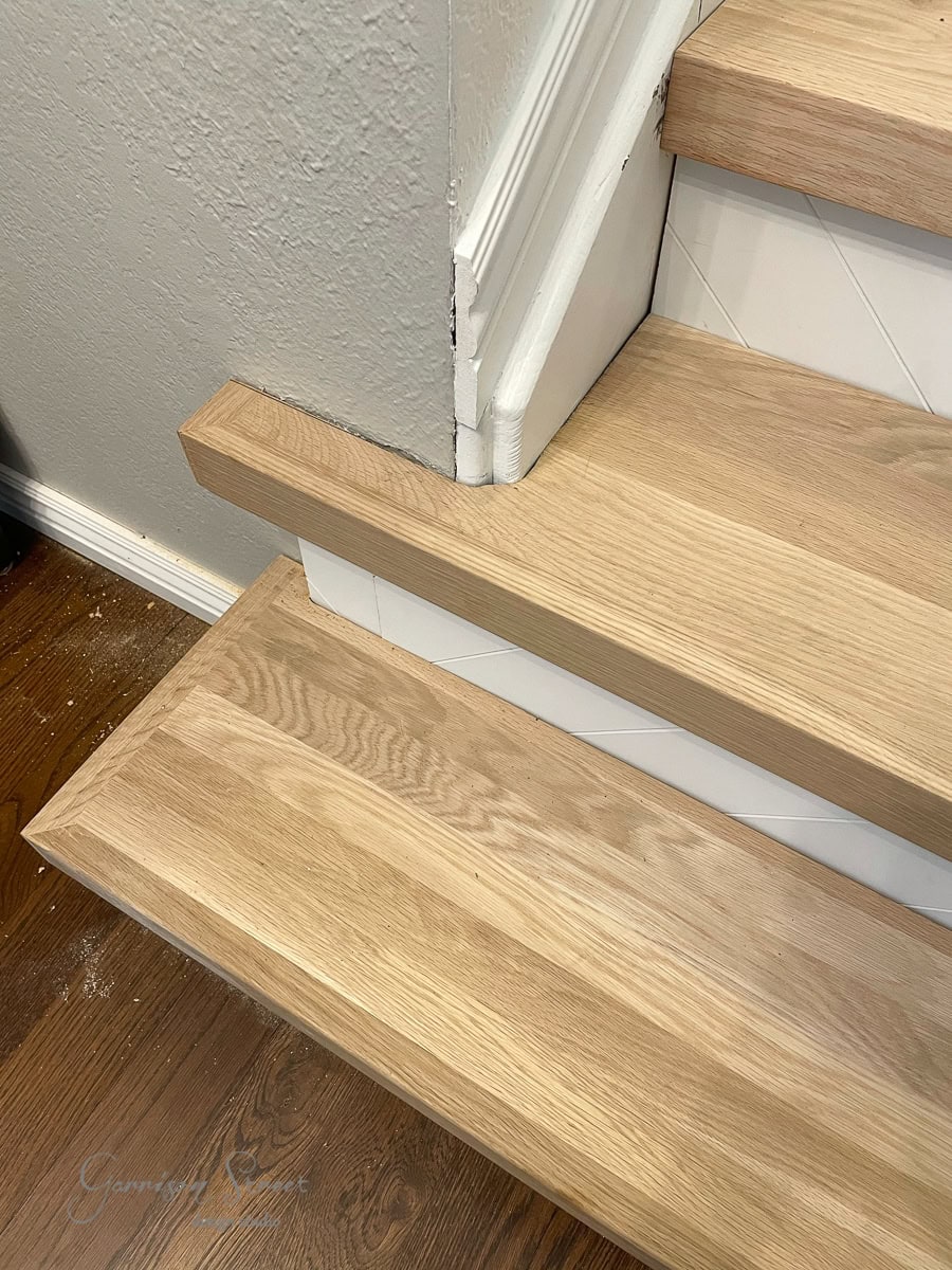A close-up of wooden stair treads with light brown steps and white risers. The corner of a wall with a textured finish is visible as the stairs meet a different type of wood flooring at the bottom.