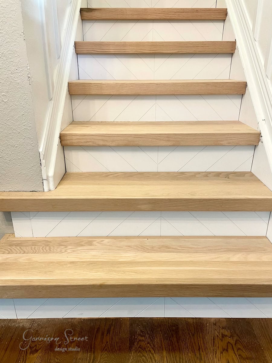 A staircase with wooden stair treads and risers features a white chevron pattern. The lower step is next to a polished wooden floor. The image is taken from the bottom looking up, while a logo in the bottom left corner reads, “Garrison Street Design Studio.”.