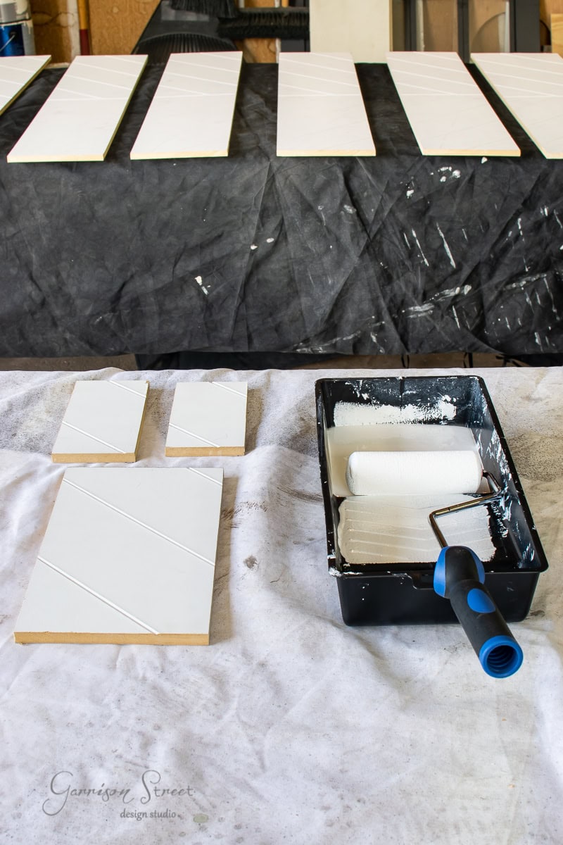 A tray with a paint roller sits on a cloth-covered surface next to painted wooden panels with diagonal grooves. More stair treads are lined up in the background, all boasting a fresh coat of white paint.