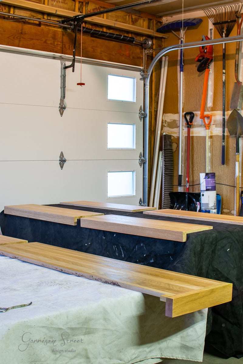 Wooden shelves and stair treads in the process of being stained are laid out on a table in a garage workshop. Various tools hang on the wall, including shovels and a rake. Sunlight filters through small windows on the garage door.