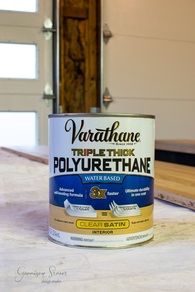 A can of Varathane Triple Thick Polyurethane sits on a wooden table, perfect for refinishing stair treads and risers. The label highlights features like an advanced self-leveling formula and clear satin finish. In the background, a garage with an open door provides the ideal setting for a DIY project.