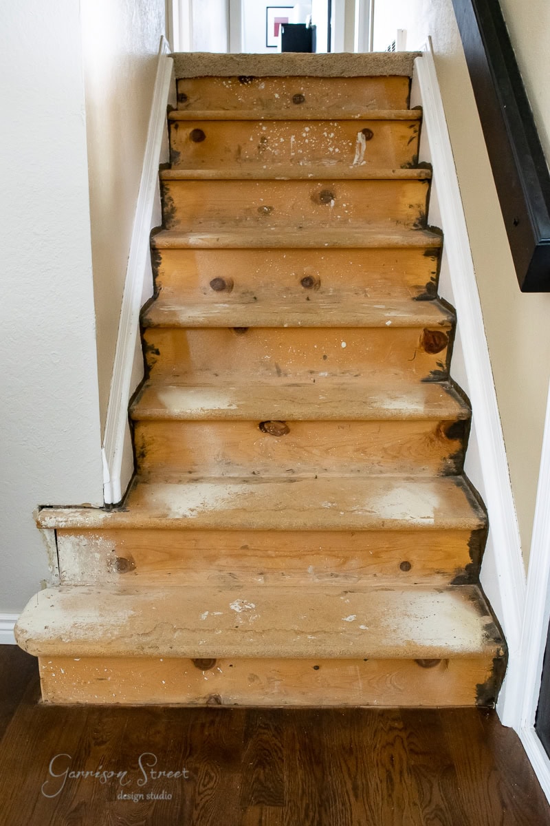 A set of wooden stairs with peeling paint and exposed wood, showing signs of wear and tear. The worn stair treads and risers lead upward, flanked by a white wall on the left and a dark handrail on the right.