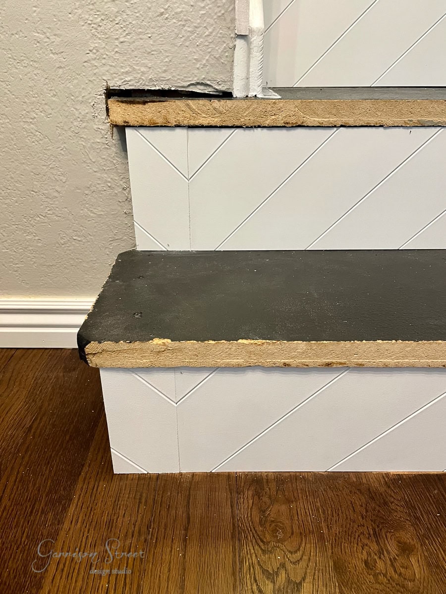 Close-up of a staircase featuring worn stairs with chipped edges and exposed layers. The white-painted risers display a herringbone pattern, contrasting against the dark stair treads. A wooden floor lies adjacent, with a baseboard running along the bottom.