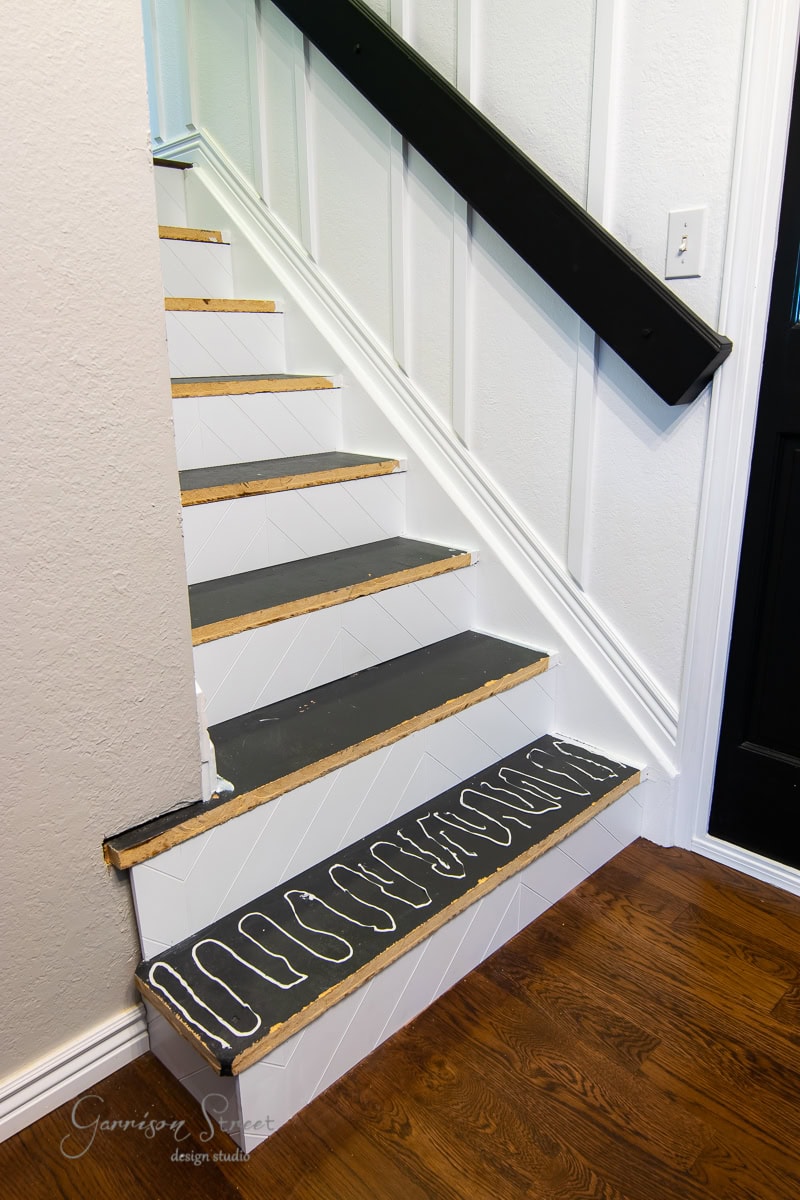 A staircase with wooden risers and painted stair treads features a stencil design on the first step. The dark handrail contrasts elegantly with the white wall, while hardwood floors spread warmly at the base.