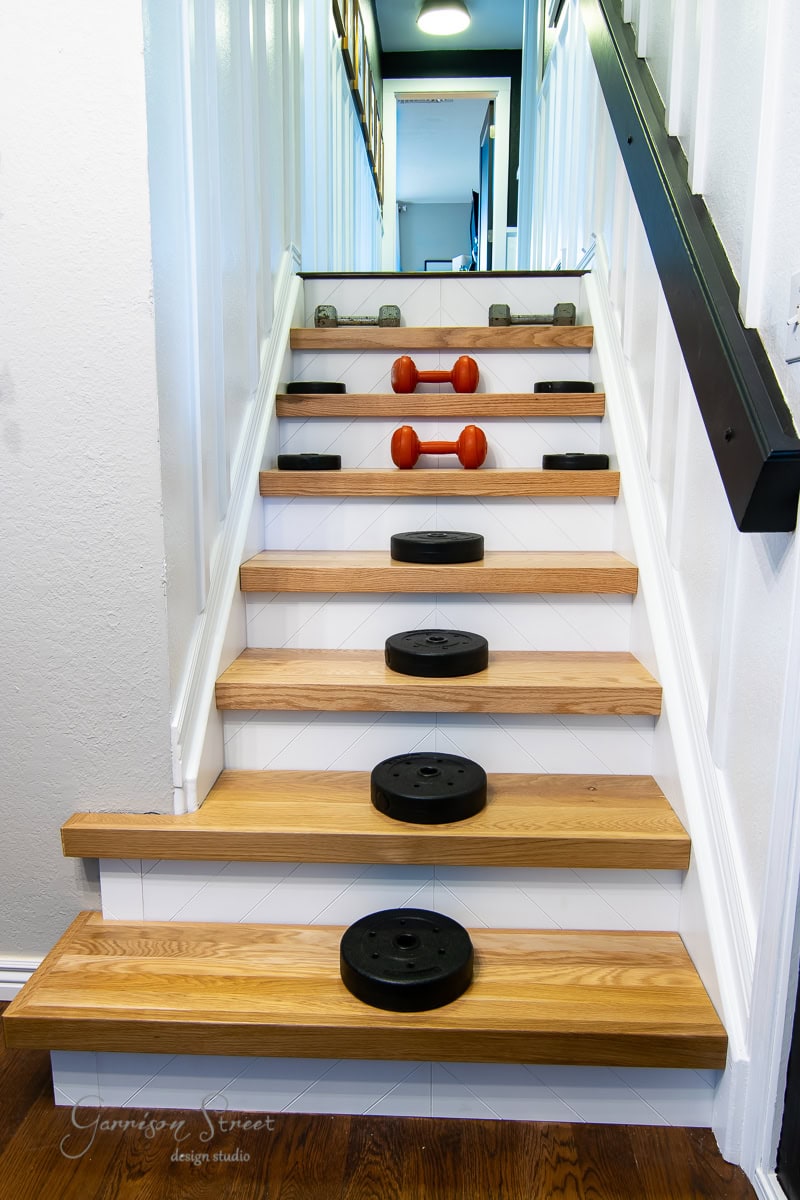 A staircase with wooden stair treads features various sized dumbbells and weight plates arranged on each step. The weights are black and orange, and the staircase is bordered by white walls and a black handrail.