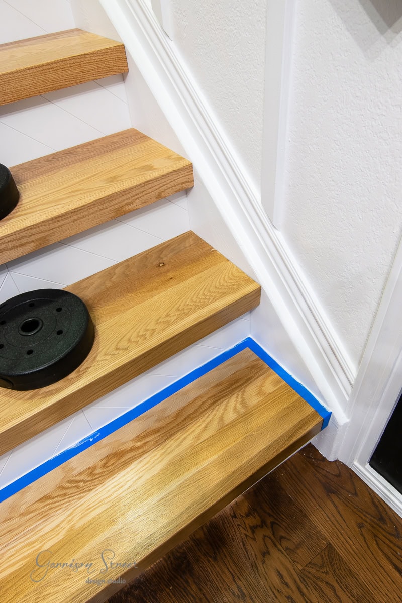 Wooden staircase with partially painted white paneling on the side. Blue painter's tape protects edges while risers and stair treads are prepped for a fresh coat. Weight plates rest on the middle step, highlighting a home improvement or renovation project in progress.