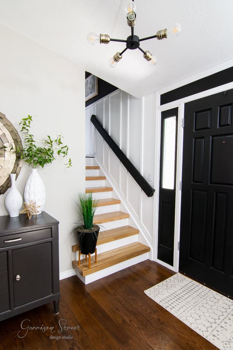 A modern entryway with a black front door, wooden staircase boasting sleek stair treads and risers, and minimal decor. A black console table with white vases and greenery sits on the left, while a round mirror hangs above. The ceiling features a contemporary light fixture.