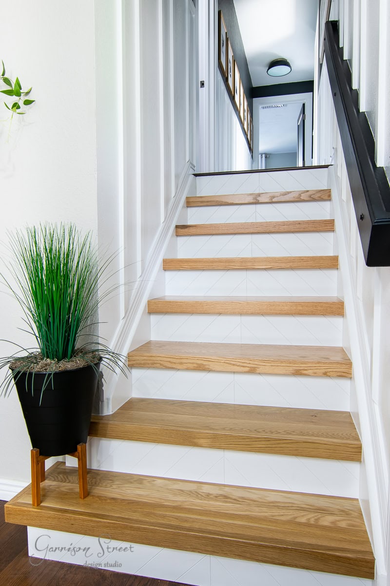 A wooden staircase features light-colored steps with matching risers leading upwards. A black handrail accompanies the journey on the right side. At the bottom left, a small green plant in a black pot adds charm, while art pieces elegantly adorn the white walls above. .