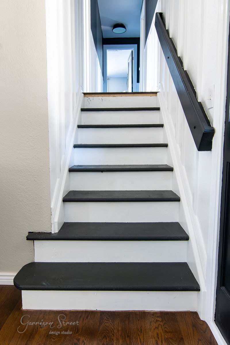 A staircase with black stair treads and white risers leads to a bright hallway. The walls are painted white, complemented by a sleek black handrail on the right side. A wooden floor graces the bottom, adding warmth and contrast to the monochrome design.