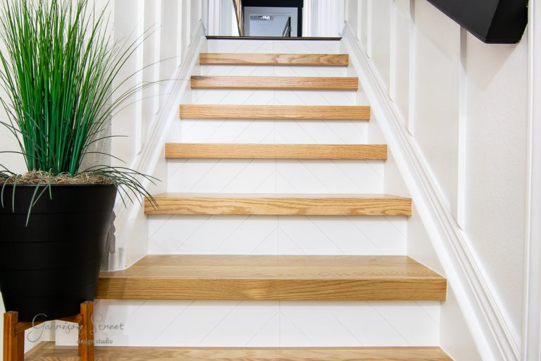A staircase with light wood stair treads and white risers ascends, flanked by pristine white walls. A potted plant with tall, thin green leaves sits on the left side. At the top, a glimpse of a room is visible.