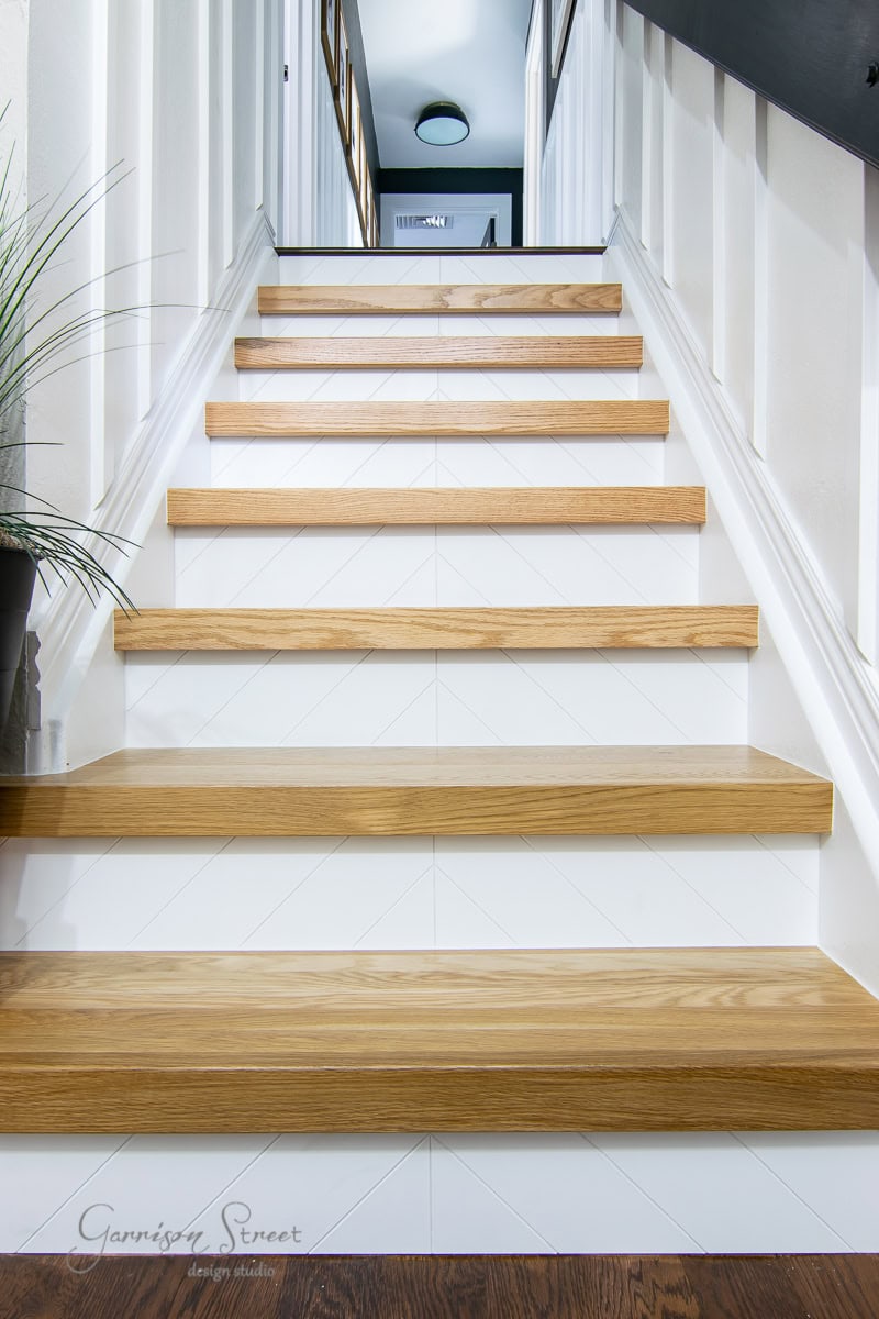 A staircase with light wood stair treads and white risers ascends between white walls. A dark handrail is visible on the right side, and a potted plant is partially visible at the bottom left. The landing at the top is out of focus.