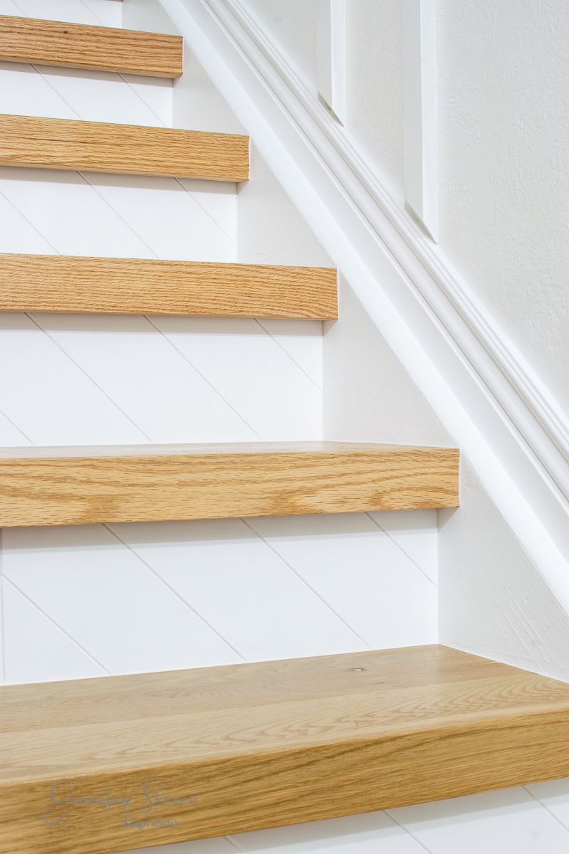 A close-up of stair treads reveals light wood paired with white risers, showcasing a subtle diagonal line pattern. The staircase exudes a minimalist and modern design, emphasizing the striking contrast between the natural wood and pristine white finish.