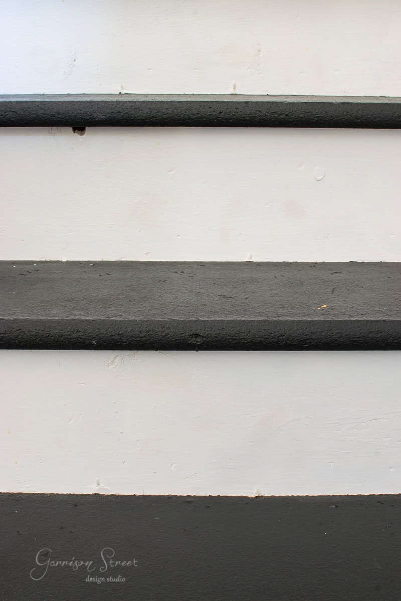 Close-up view of three stair steps featuring dark gray stair treads and white risers. The minimalist and modern design complements the space beautifully. The logo "Garrison Street Design Studio" appears in the lower left corner.