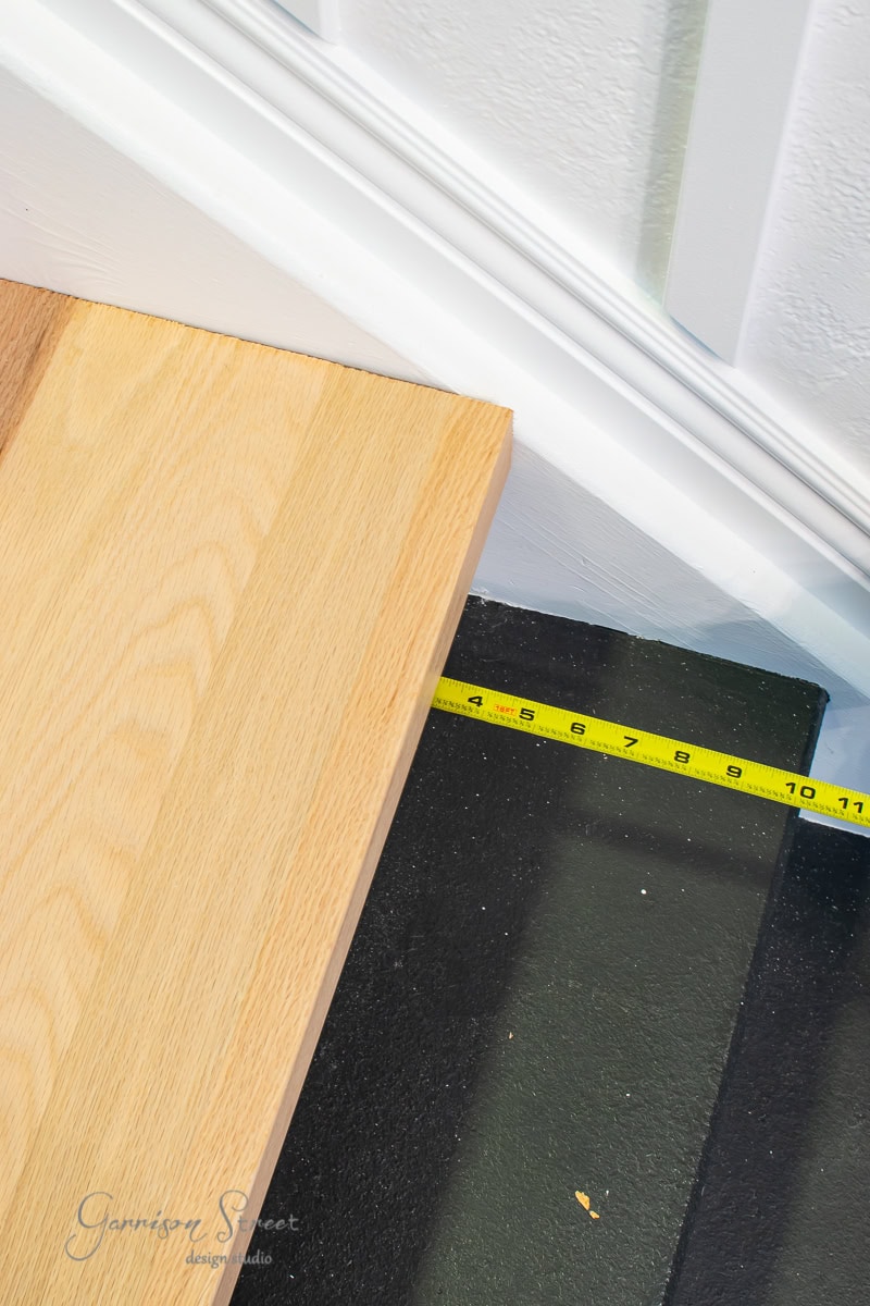 A yellow measuring tape measures the width of a wooden plank against a white wall, reminiscent of risers aligning with the stair treads. The floor beneath is dark, contrasting with the light wood and wall.