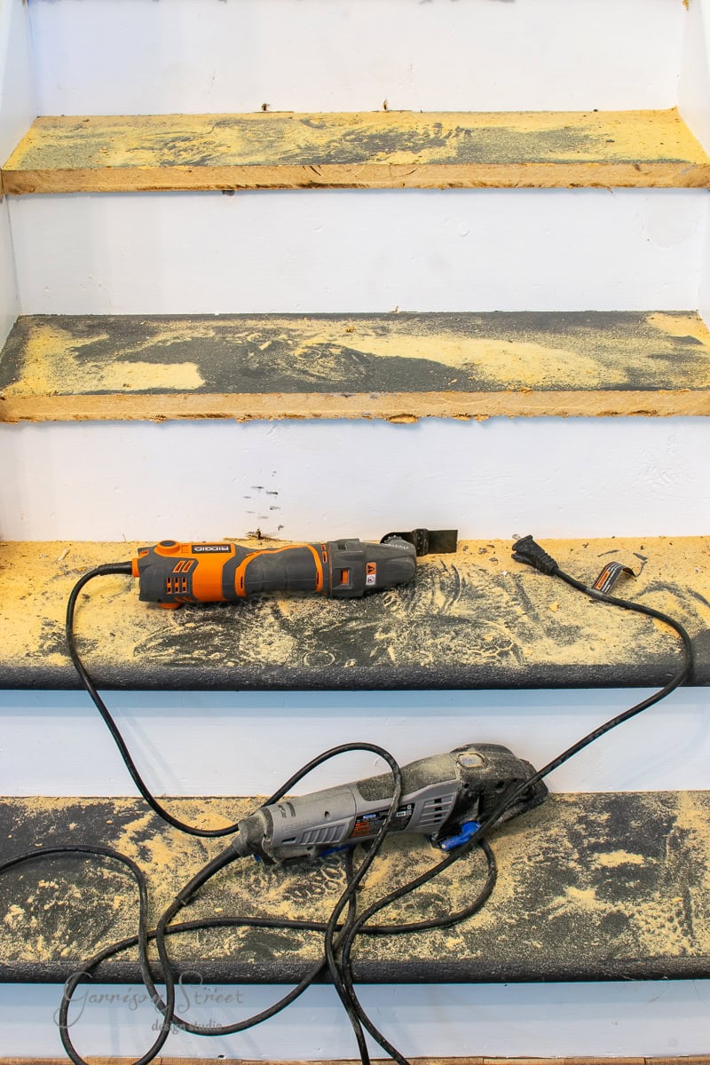 A set of wooden stairs, their risers and stair treads dusted with sawdust, reflect ongoing carpentry work. Two corded power tools rest on the steps, hinting at renovation. The white stairs show clear signs of sanding and wear.