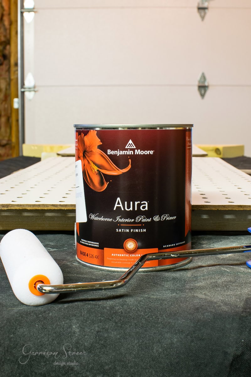 A can of Benjamin Moore Aura waterborne interior paint in satin finish is on a table, ready to transform your kitchen island or cabinet. The can, adorned with a floral design, sits beside a paint roller, with the backdrop of a perforated board and part of a garage door.