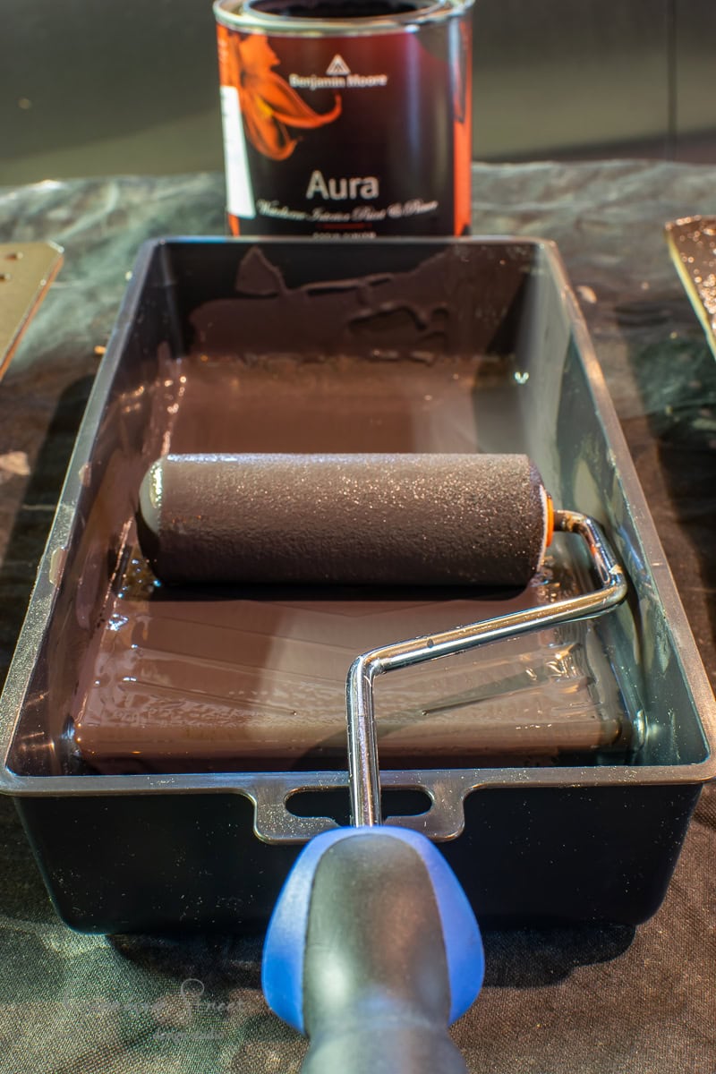 A paint roller with a blue handle rests in a black paint tray filled with black paint. In the background, there is a can of Benjamin Moore Aura paint sitting on a black drop cloth, ready to transform cabinets or refresh the look of your kitchen island.