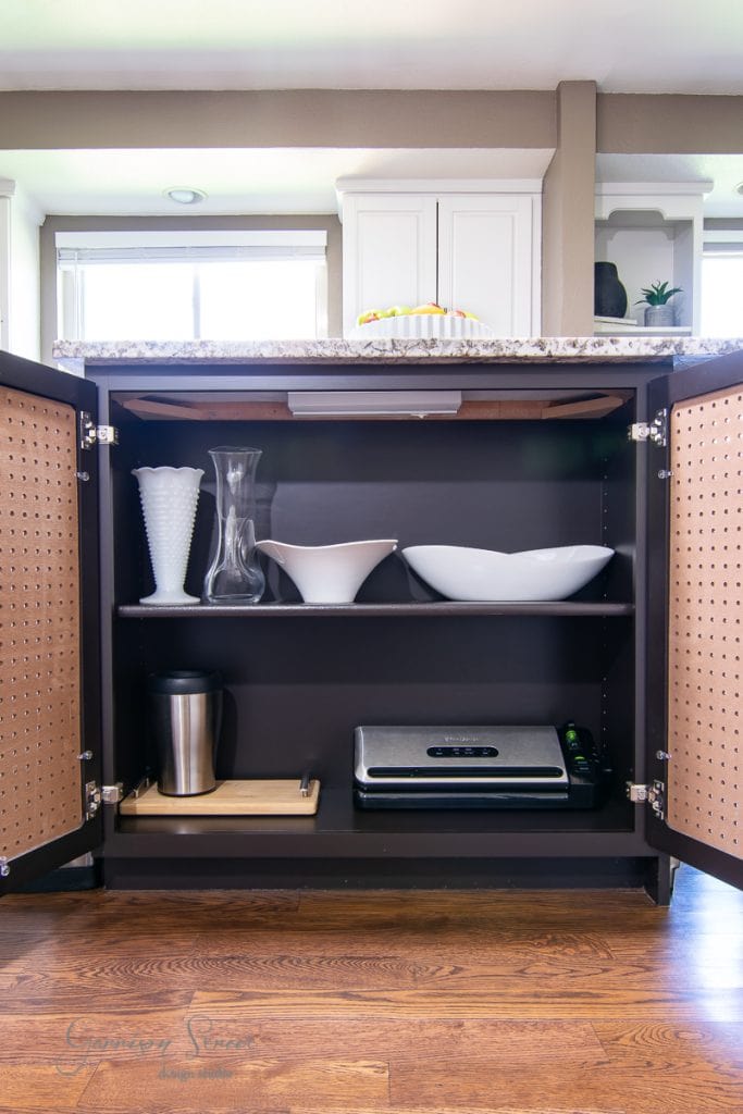 Open kitchen cabinet with wooden shelves. Upper shelf holds white ceramic bowls and vases. Lower shelf contains a silver kitchen appliance and a trash bin. The cabinet, adjacent to the granite-counter-topped kitchen island, has pegboard doors and is bathed in bright natural light from windows.