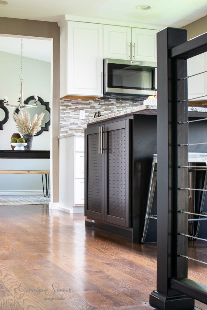 Modern kitchen with dark cabinets and light countertops, complemented by a sleek kitchen island. A microwave is mounted above the stove, while the backsplash showcases a geometric tile pattern. A dining area with a chandelier is visible in the background, and the floor is polished wood.