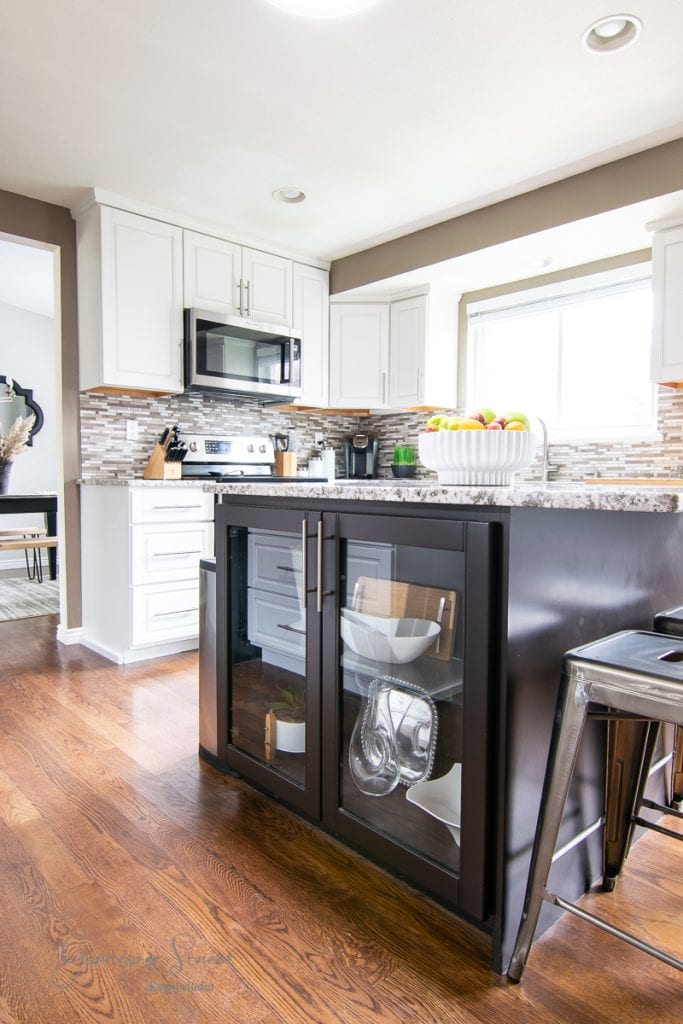 A modern kitchen features white cabinets and a tiled backsplash with stainless steel appliances. The black kitchen island, adorned with glass-front cabinets displaying dishware, is complemented by bar stools tucked underneath and a bowl of fresh fruit gracing the countertop.