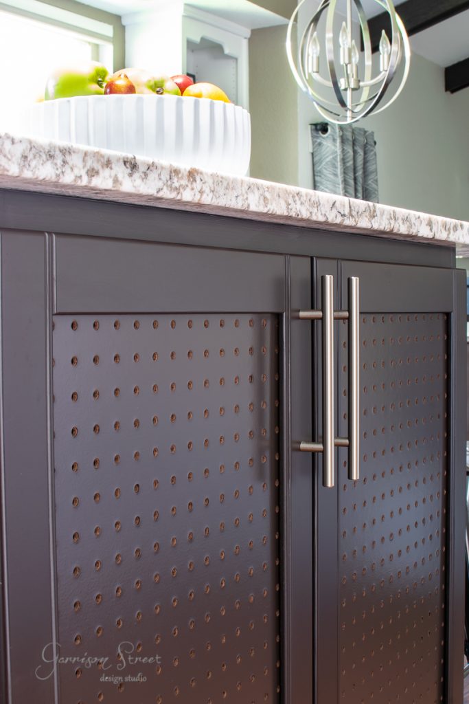 A close-up of dark brown kitchen cabinets with textured panels and sleek silver handles frames the elegant kitchen island. The marble countertop holds a large white bowl filled with assorted fruits, while a modern light fixture hangs above, casting a warm glow over the scene.