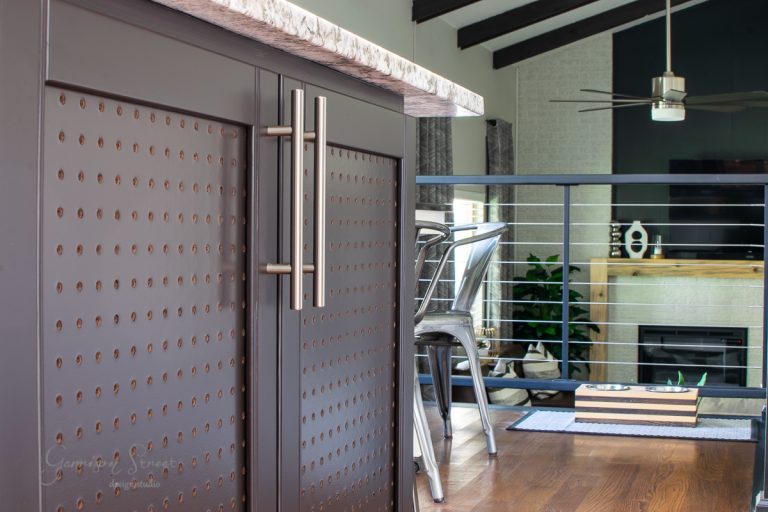 Close-up of a kitchen island featuring dark cabinetry with modern metallic handles. The marble countertop gleams under the light. In the background, metal chairs surround a fireplace and TV in the living area. A ceiling fan spins gently from the exposed beam ceiling.