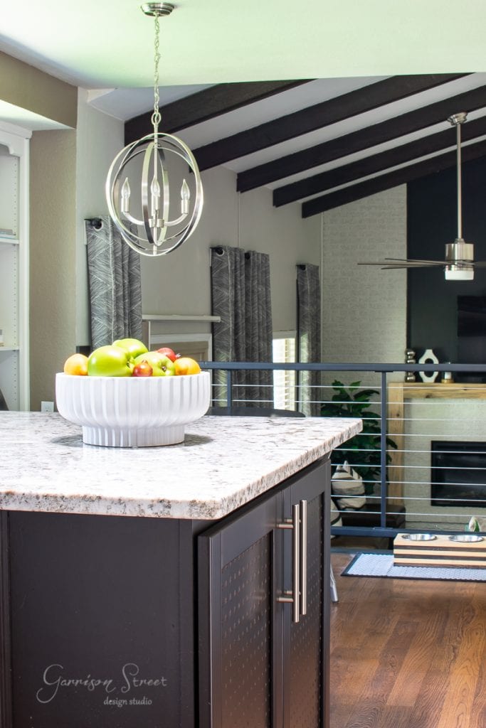 Modern kitchen and living area with dark wood beams overhead. A large, stylish light fixture illuminates the granite kitchen island, which features sleek cabinets and holds a white bowl filled with colorful apples. Grey curtains and a metal railing complete the look.