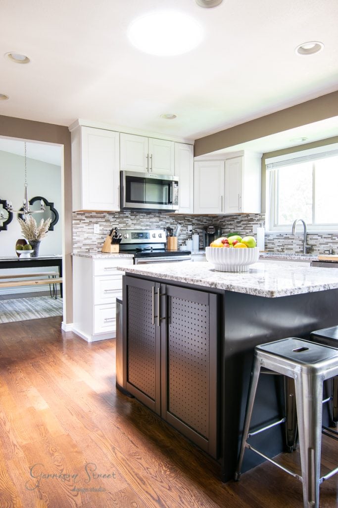 The modern kitchen boasts sleek white cabinets and a stainless steel stove, complemented by a microwave. A granite-topped kitchen island with a black base is surrounded by metal bar stools, adorned with a fruit bowl. Wood flooring extends into the dining area, while natural light fills the space.