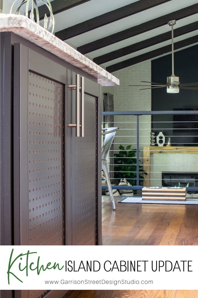 A close-up of a sleek kitchen island with dark cabinetry and a speckled countertop. The backdrop reveals a living area featuring a table, chairs, and a ceiling fan. Large windows and a fireplace accentuate the bright and contemporary space.