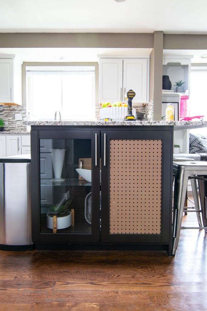 The sleek black kitchen island boasts a granite countertop and glass cabinet doors, perfectly showcasing organized pots and pans. A tool rests on the counter, while pegboard detail adds charm. Modern white cabinetry, barstools, and a stainless steel trash can complete the elegant space.