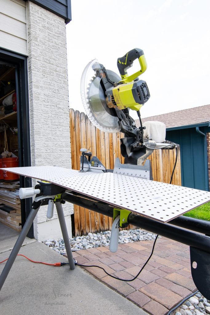 A miter saw is positioned on a saw stand outside near a garage, ready to transform a white perforated panel, perhaps destined for a kitchen island feature. A wooden fence and part of a building frame the scene, with rocks and a grassy area nestled at the base.