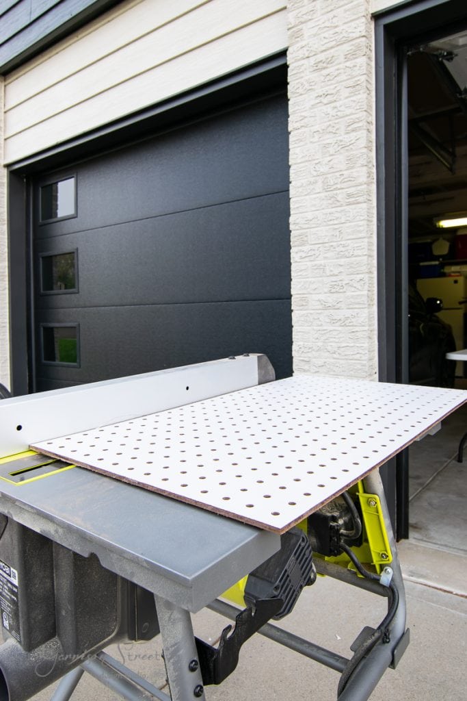 A white perforated panel is placed on a table saw outdoors near an open garage, possibly for customizing a kitchen island. The saw, with its bright green base, rests on a concrete surface, while a black garage door is partially visible in the background.
