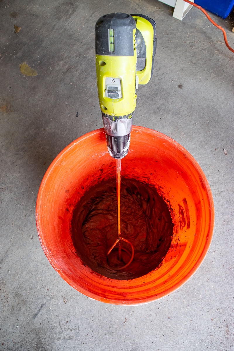 A drill with a mixing attachment hovers above a large orange bucket, stirring a dark substance inside. Resting on a concrete floor.