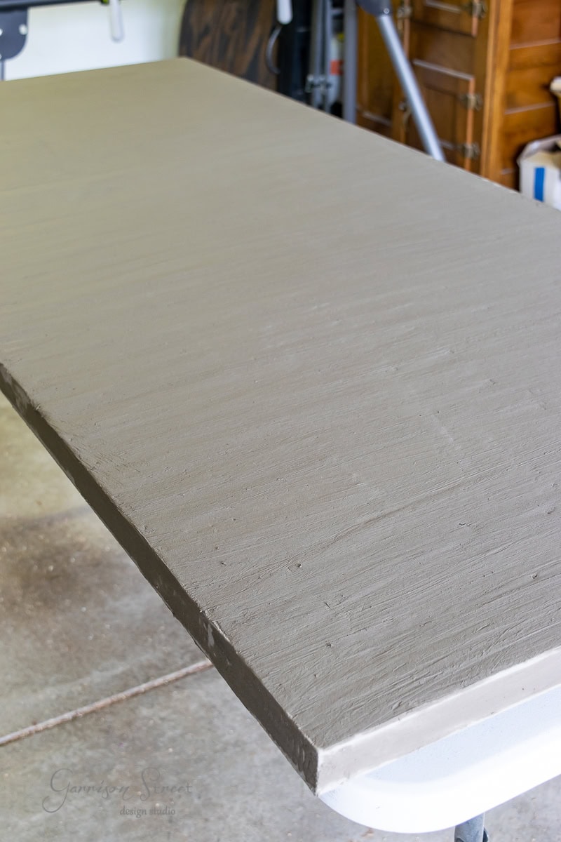 A DIY concrete outdoor table with a smooth, gray surface is showcased in a room filled with various tools and furnishings in the background. The rectangular table boasts a sturdy build and a simple, modern design ideal for any setting.