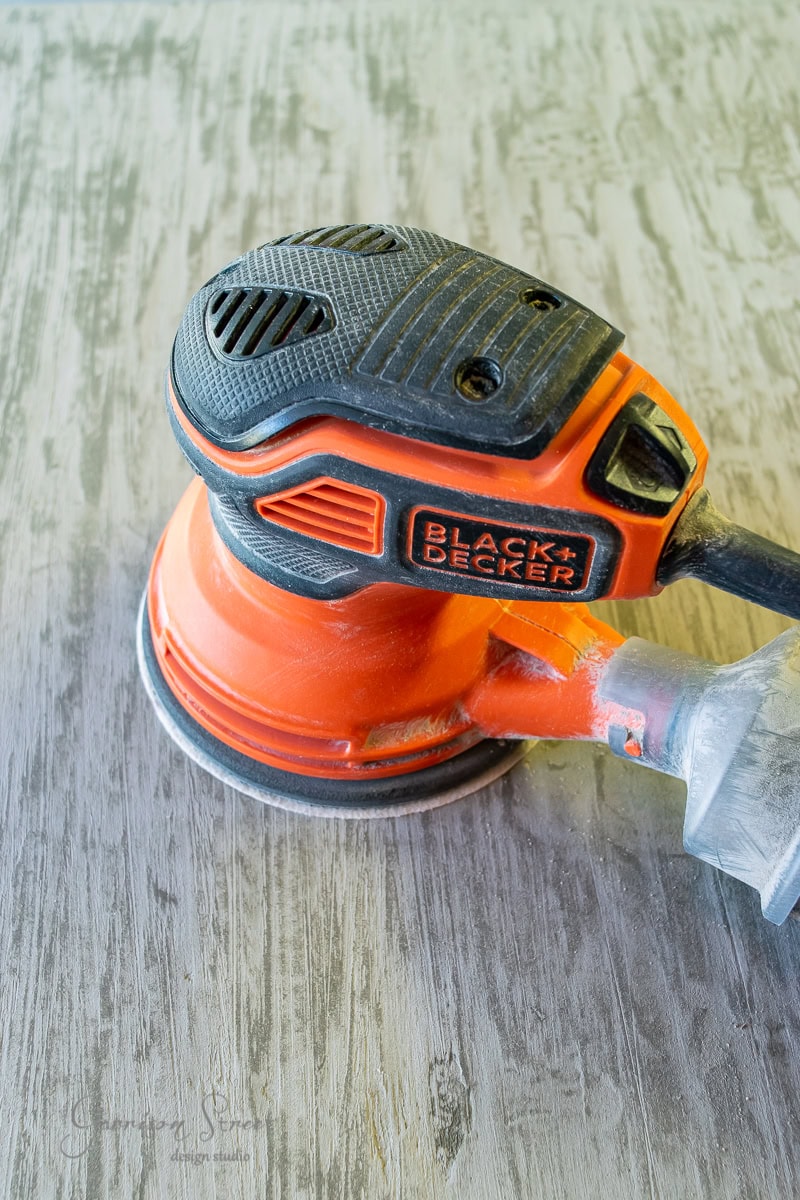 A Black+Decker orange and black electric sander lies on a textured wooden surface. It's plugged in and resting, with visible dust from recent sanding activity covering the wood.