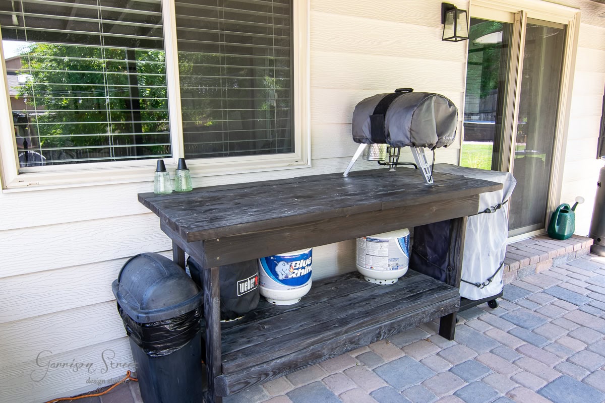 A wooden outdoor table with a small covered grill, propane tank, and two covered bottles on top. Below are large propane and water tanks. A window and sliding door complete the scene on the brick patio.