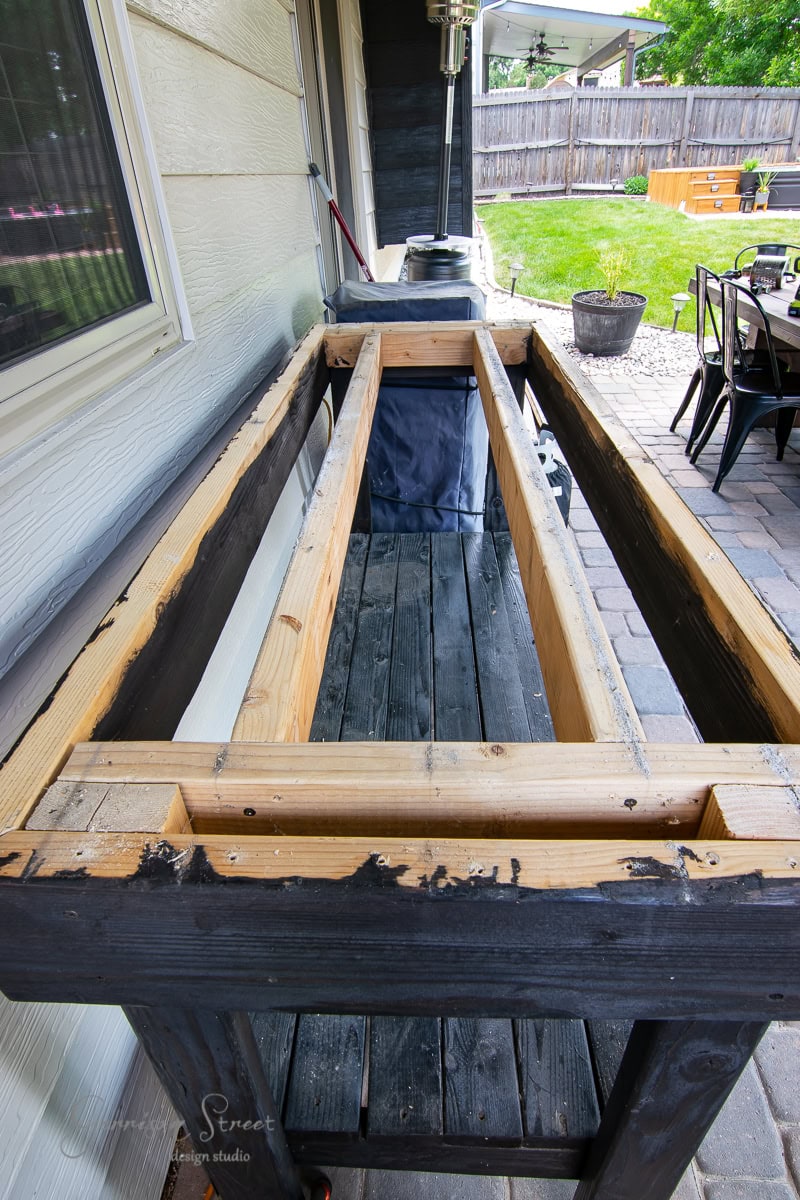 A wooden frame with an open top sits on the brick-paved patio next to a house. Patio furniture and a grill are visible nearby, adding character to the inviting outdoor space.