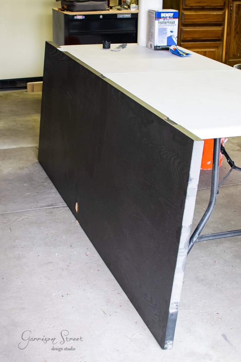 A large blackboard is propped up against a white folding table in the garage. The table holds a container of foam sealer and various tools. Nearby, wooden cabinets and a toolbox provide easy access to essential equipment.