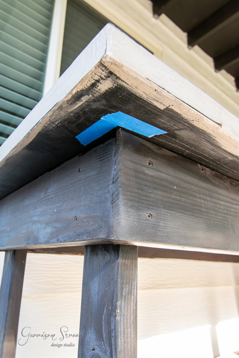 Close-up of a wooden table corner with a sleek concrete top, perfect for a DIY outdoor setup. The underside reveals a piece of blue tape, likely used during construction or finishing. Sunlight beautifully highlights the wood's natural texture.