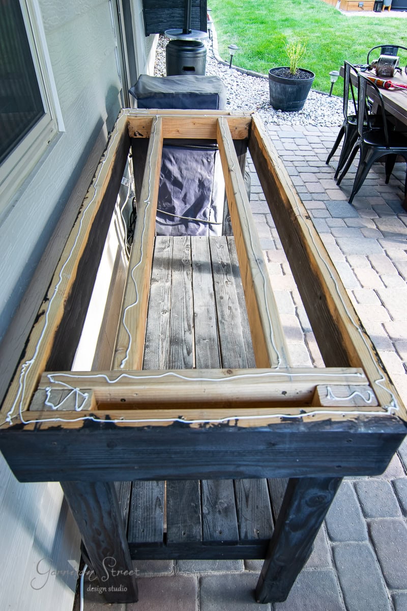 A wooden outdoor planter box on a patio, partially assembled with a black frame and natural wood slats, complements the DIY concrete outdoor table nearby. Coils of water-resistant caulking are applied along the edges. In the background, there's a dining table set and a neatly maintained lawn.
