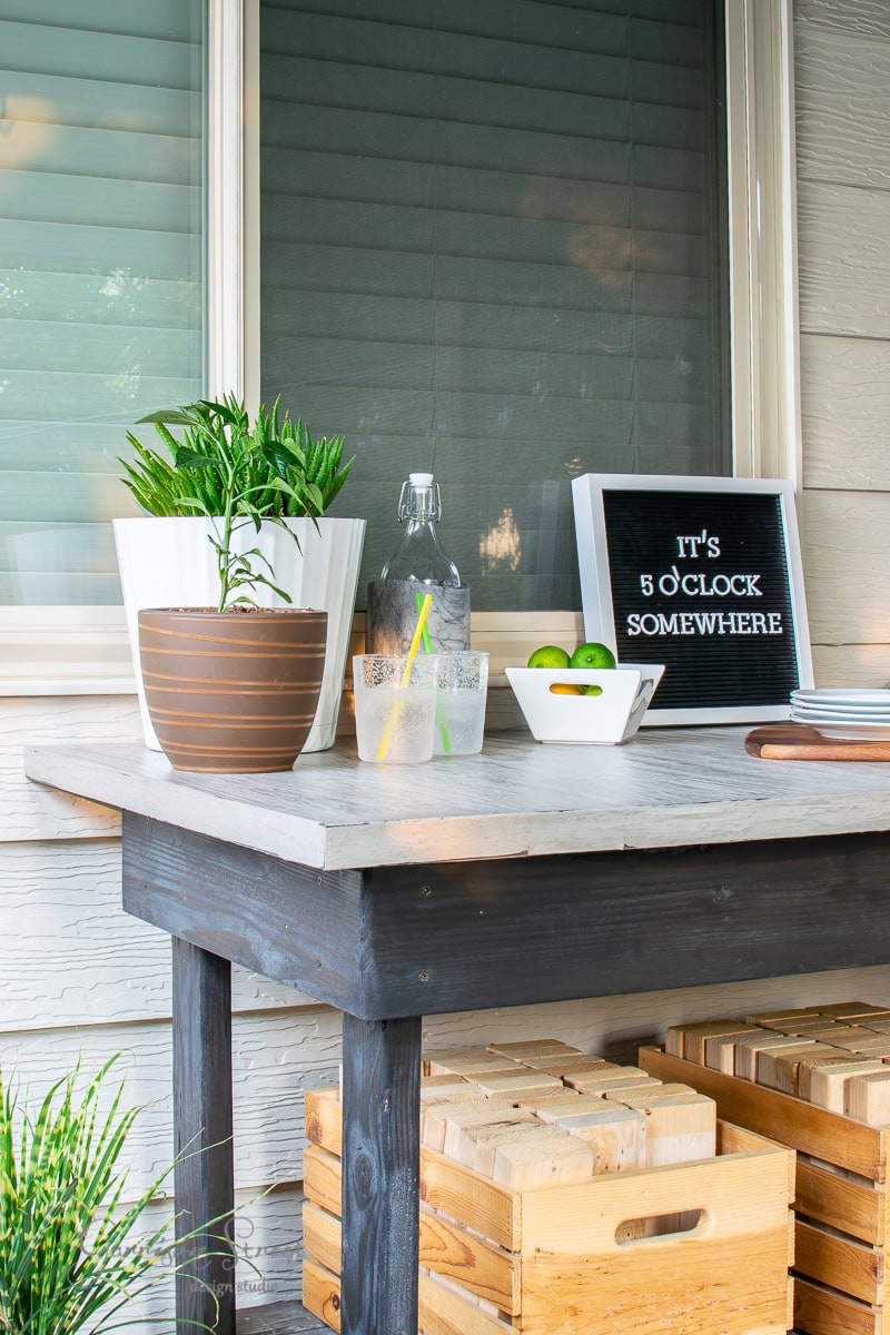A DIY concrete outdoor table hosts potted plants, a bowl of limes, two drinks with straws, and a letter board reading "It's 5 o'clock somewhere." A stack of plates and a bottle are also on the table. The setting is relaxed and inviting.