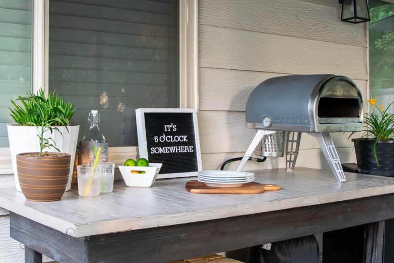 A DIY concrete outdoor table holds a pizza oven, plates, a cutting board, and drinks. A letter board reads "It's 5 o'clock somewhere." The table is adorned with potted plants and a bottle with glasses, creating the perfect backyard retreat.