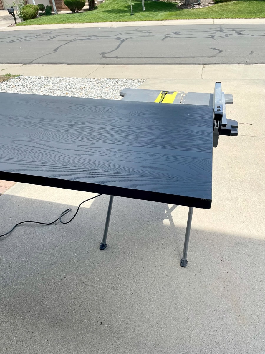 A large, rectangular black wooden board rests on a saw table in a driveway. The wood grain texture contrasts with the small pebbles and street visible in the background.