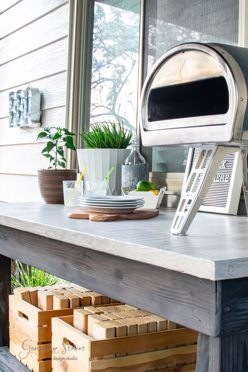 A charming outdoor kitchen setup showcases a DIY concrete table elegantly supporting a pizza oven. Nearby, potted plants and glasses with straws complement the scene, while a small cutting board with a lemon adds zest. Crates tucked beneath complete this inviting space with trees reflecting in the window.