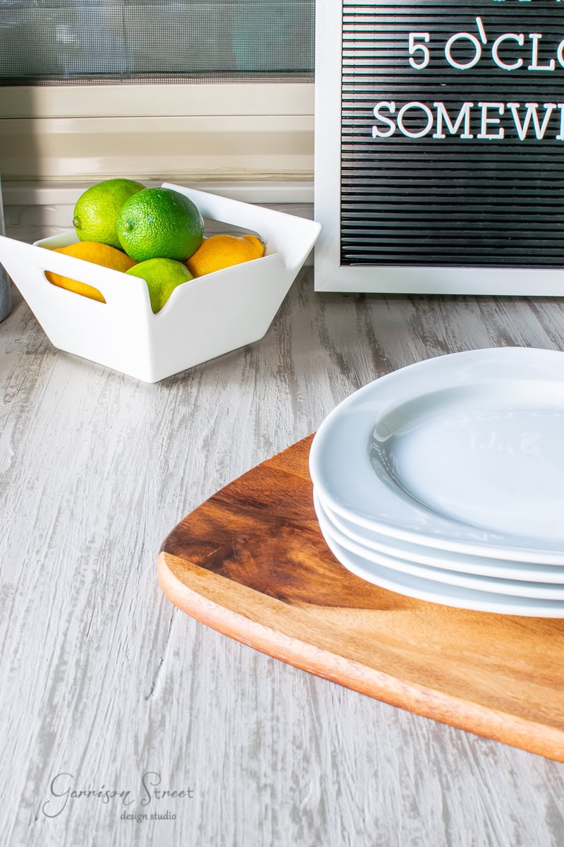 A stack of white plates on a wooden board, with a ceramic bowl of lemons and limes beside them, sits elegantly on the concrete tabletop. In the background, a letter board reads "5 o'clock somewhere," enhancing the laid-back vibe on this light wooden surface.