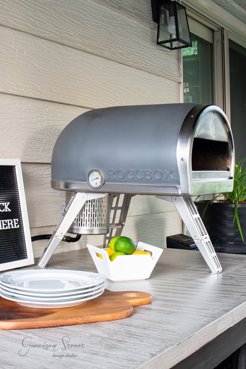 A portable Roccbox pizza oven sits on the concrete tabletop on the patio, beside a cutting board with white plates. A small bowl with limes and a decorative plant are near the oven. A sign partially visible reads, "...CK HERE," complementing the casual outdoor setting.