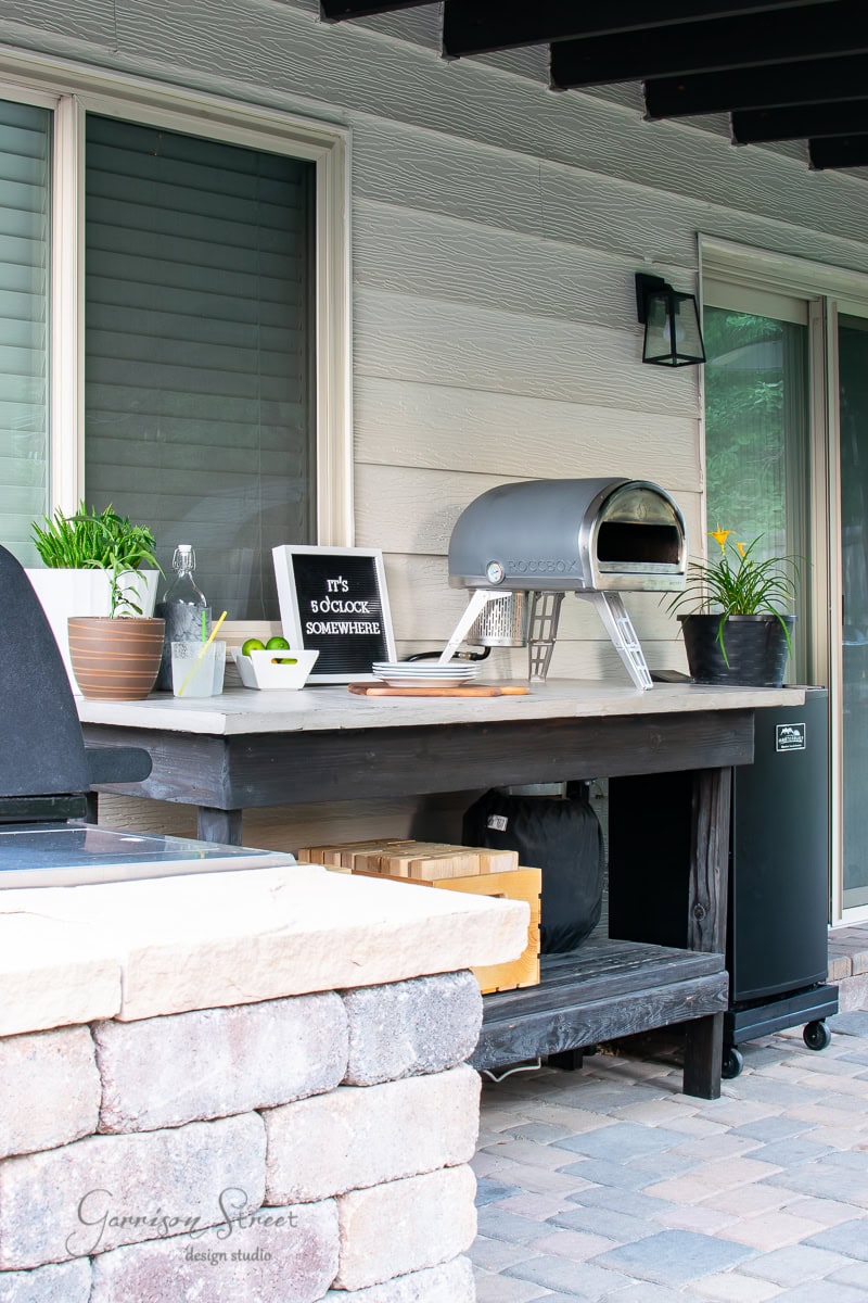 An inviting outdoor patio setup features a concrete-topped table, complemented by a grill and pizza oven. Potted plants add a touch of green, while a decorative sign humorously reads "It's 5 o'clock somewhere" against the light-colored wall and large window backdrop.